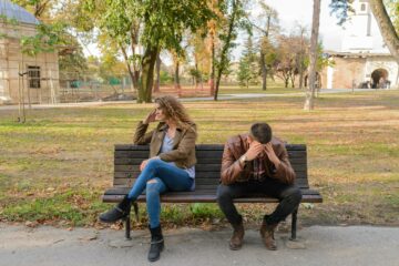 A man and woman are sitting on a bench; the woman is looking away with her hand to her head; the man has his head in his hands.