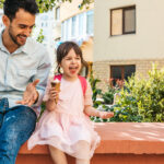 Image of happy cute little girl sitting with dad on the city street and eating ice-cream outdoor. Fun girl kid and father have fun and playing outside.