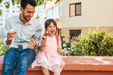 Image of happy cute little girl sitting with dad on the city street and eating ice-cream outdoor. Fun girl kid and father have fun and playing outside.